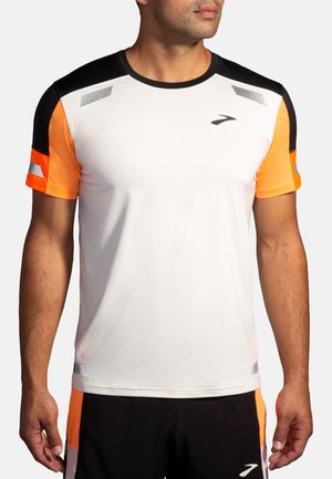 Hombre con camiseta deportiva blanca con mangas negras y naranjas, detalles reflectantes y pantalones cortos a juego en negro y naranja.