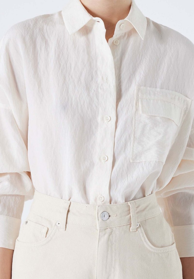 Personne portant une chemise texturée blanche à boutons rentrée dans un pantalon taille haute beige clair avec un bouton et des passants pour ceinture.