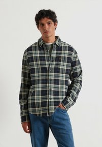 Camisa de cuadros con un patrón de cuadros en azul marino y verde claro. Hecha de una tela suave, tiene un cierre de botones en el frente y un collar en punta.