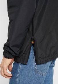 Personne portant une veste noire avec une fermeture éclair latérale partiellement ouverte sur un jean bleu, montrant un poignet élastique et une main détendue sur le côté.