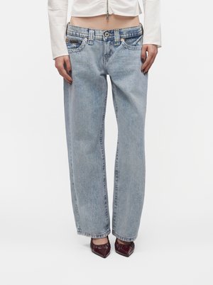 Kvinde iført lyseblå højttaljede, lige jeans, hvid crop top med lange ærmer og mørkerøde spidse sko, stående på hvid baggrund.
