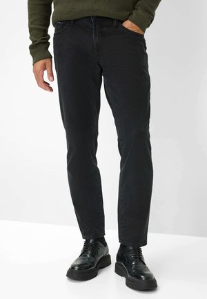 Schwarze Denim-Jeans mit schmalem Schnitt und subtiler Struktur. Kombiniert mit schwarzen Schuhen, die über kunstvolle Details und eine dicke Sohle verfügen.