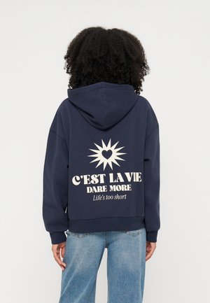 Personne portant un sweat à capuche bleu marine avec un motif soleil et cœur et le texte "C’EST LA VIE DARE MORE Life’s too short" au dos.