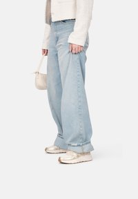 Jeans a gamba larga blu chiaro in denim, con orlo sfilacciato. Indossati con sneakers argento metallizzato e portando una piccola borsa beige.