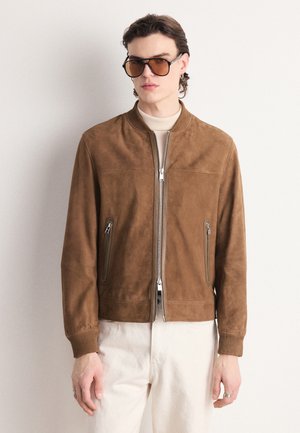Veste bomber en daim marron avec des poignets et un ourlet côtelés, dotée d'une fermeture éclair centrale et de deux poches latérales. Le mannequin porte des lunettes de soleil teintées.