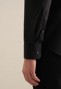 Zwarte lange mouwen shirt met een knoopmanchet met een hexagonaal logo-detail en contrasterende knopen tegen een gladde stofstructuur.