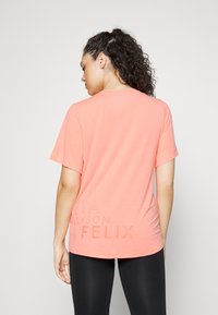 T-shirt atlético leve, em coral, com mangas curtas, decote redondo e detalhes em texto na parte inferior das costas. Combinado com leggings pretas.