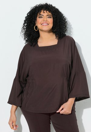 Mujer sonriente con cabello rizado y negro que lleva pendientes de aro dorados, una blusa suelta marrón oscuro con mangas anchas y pantalones a juego.