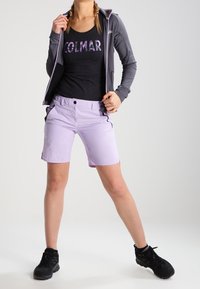 Mujer vestida con una camiseta de tirantes negra de Colmar, una chaqueta gris con cremallera, pantalones cortos lila claro y zapatillas negras, de pie con las piernas separadas sobre un fondo blanco.