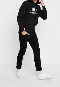 Czarna bluza z kapturem z przednią kieszenią i białym logo "ALPHA INDUSTRIES", w połączeniu z czarnymi dżinsami slim fit i białymi sneakersami.