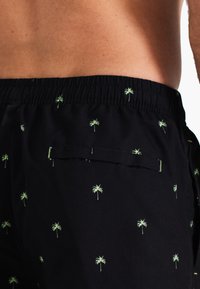 Zwarte zwemshorts van lichtgewicht stof, met kleine groene palmboomprints en een elastische tailleband met een achterzak.