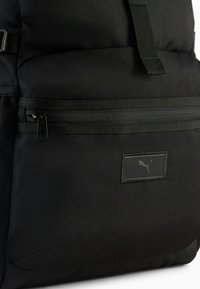 Sort rygsæk med struktureret stof, en lynlåslomme på fronten og et mærketag med et logo. Inkluderer justerbare stropper og et slankt design.