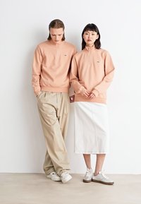 Felpe a zip pesca indossate con pantaloni beige e una gonna bianca. Il tessuto sembra liscio con una vestibilità comoda. Le scarpe sono grigie chiare con dettagli.
