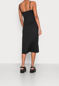 Mujer vistiendo un vestido midi negro de tirantes delgados y sandalias de plataforma negras, de pie frente a un fondo claro y liso.