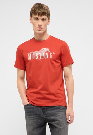 Junger Mann mit kurzem blondem Haar, der ein rotes Mustang-T-Shirt und dunkelgraue Jeans trägt, steht mit einer Hand in der Tasche.