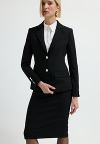 Blazer nero su misura con bottoni dorati e due tasche frontali, abbinato a una gonna a tubino coordinata. Sotto indossa una camicia bianca con colletto.