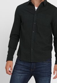 Dunkelgrünes Hemd mit Knöpfen aus weichem Stoff, langen Ärmeln, einer Brusttasche und abgerundetem Saum. Kombiniert mit blauen Jeans.