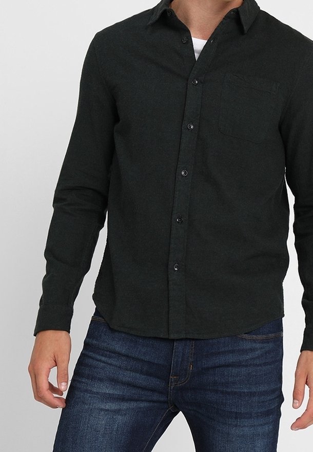 Dunkelgrünes Hemd mit Knöpfen aus weichem Stoff, langen Ärmeln, einer Brusttasche und abgerundetem Saum. Kombiniert mit blauen Jeans.