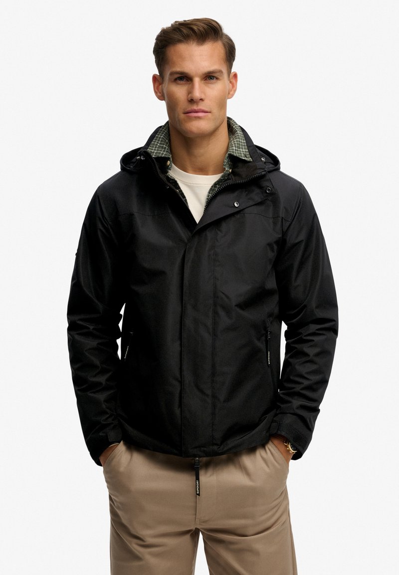 Black waterproof jacket with a hood, featuring a patterned green collar, zippered pockets, and smooth fabric. Worn over a gray shirt.