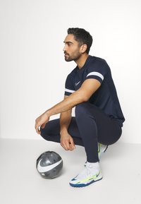 Marine sports t-shirt med hvide striber, marine track pants og hvid/blå fodboldsko. En sort og sølv fodbold ligger foran.