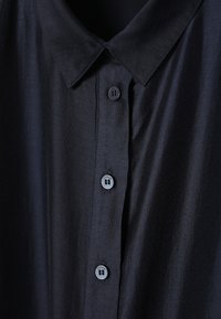 Devant de chemise en satin noir avec col pointu et boutons visibles à quatre trous sur la patte de boutonnage.