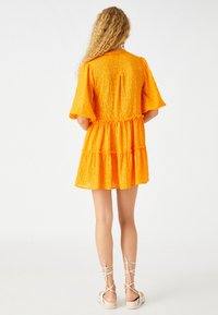 Helles oranges Kleid mit kurzen, ausgestellten Ärmeln, strukturiertem Stoff und einem gestuften Rockdesign. Getragen mit beige, schnüren Sandalen.