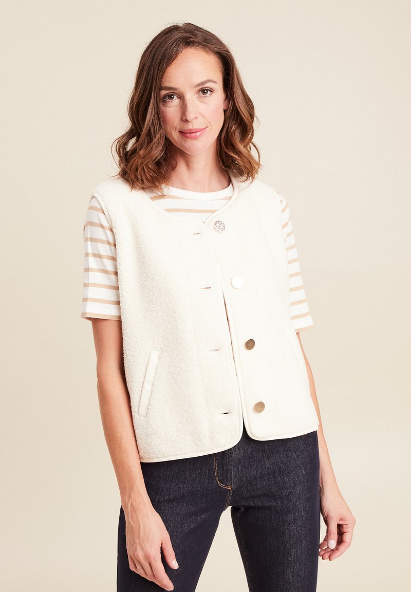 Weißer Fleece-Weste mit Rundhalsausschnitt, vorderer Knopfleiste und Seitentaschen, über einem gestreiften Kurzarmhemd getragen. Dunkle Jeans sind sichtbar.