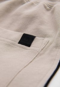Jogger beige in tessuto con etichetta nera nella tasca, caratterizzato da una texture liscia e una vita elasticizzata. Gli accenti includono bordi profilati in contrasto.