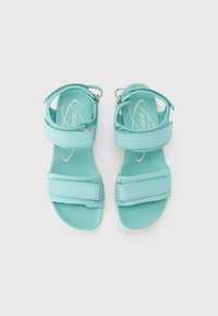 Τυρκουάζ σανδάλια με δύο φαρδιές λωρίδες, μαλακό υλικό, ρυθμιζόμενη λουρί στον αστράγαλο και λευκή σόλα. Ο λογότυπος της μάρκας φαίνεται στις σόλες.