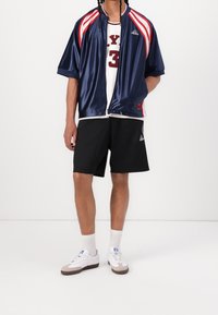 Námořnická saténová bunda na zip s červenými a bílými pruhy, bílý bezrukávový tričko s červeným nápisem, černé šortky, bílé tenisky s šedými detaily.