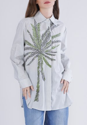 Mujer con camisa de botones de rayas gris claro con una gran palmera bordada en verde y negro en el frente, combinada con jeans azules.