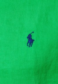 Grön linneväv med en liten, mörkblå broderad polo-spelare-logga. Slät textur med en enkel, enfärgad design.