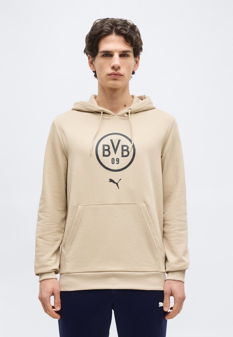 Junger Mann trägt beige BVB 09 Hoodie mit Fronttasche und schwarzem Puma-Logo, steht vor einfachem weißem Hintergrund.