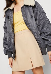 Chaqueta bomber gris con cuello de piel sintética, combinada con una blusa amarilla de canalé y una falda beige con una abertura frontal.