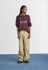 Bordo majica s dugimi rukavi in teal dodatki ter logotipom "NIKE"; bež cargo hlače s žepi in širokimi nogami; črne superge z rumenimi detajli.