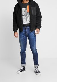 Sort fleecejakke med stor hætte, lagdelt over en grafisk sort T-shirt. Slim-fit blå jeans og sorte høj-til-top sneakers med hvide snørebånd.