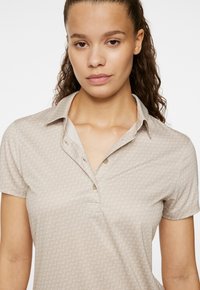 Polo beige con un sottile motivo decorativo, dotato di bottone e maniche corte. Il materiale è morbido e leggero.