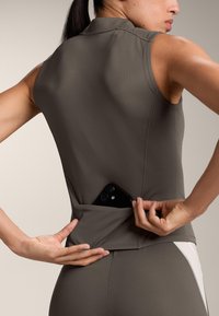 Vrouw in grijze mouwloze top steekt smartphone in klein achterzakje, gezien van achteren met focus op zak en handen.