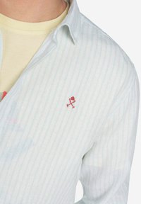 Camisa a rayas en azul claro y blanco, con cuello y un acento bordado en rojo en el lado izquierdo. Textura de tela suave.
