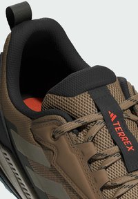 Brązowe siatkowe buty trekkingowe z czarnym wyściełanym kołnierzem i czerwonymi akcentami, z szarymi nakładkami i teksturowanymi sznurowadłami dla zwiększonej trwałości.