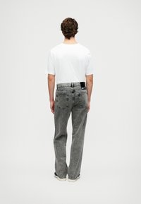 Hombre de pie de espaldas usando una camiseta blanca, vaqueros gris desgastados y zapatillas blancas contra un fondo liso.