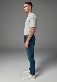Tricou gri deschis cu mânecă scurtă, blugi din denim albaștri și adidași albi. Blugii au un stil tradițional cu cinci buzunare. Modelul stă de profil.