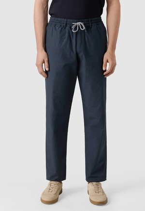 Uomo che indossa pantaloni casual blu scuro con coulisse e sneaker beige, in piedi contro uno sfondo chiaro e uniforme.