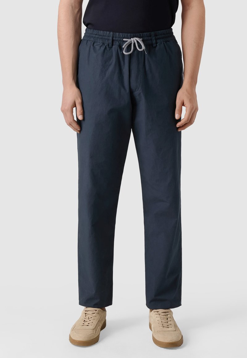 Mann trägt dunkelblaue, lässige Hose mit Kordelzug und beige Turnschuhe, steht vor einem schlichten, hellen Hintergrund.