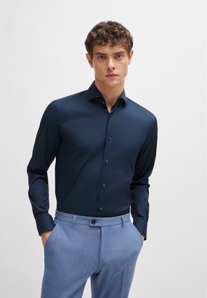 Cămașă albastru închis cu mânecă lungă dintr-un material neted, cu guler clasic și închidere cu nasturi pe față, combinată cu pantaloni tailored de culoare albastru deschis.