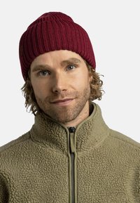 Red ribbed beanie made of knitted fabric, featuring a textured design. Paired with a light olive fleece jacket with a zippered collar.