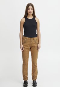 Haut noir sans manches associé à un pantalon slim beige avec des coutures décoratives, une fermeture à cinq boutons et des poches latérales. Chaussures noires.