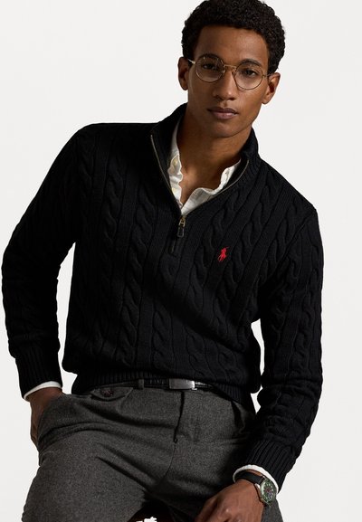 Maglione nero a lavorazione a trecce con colletto a mezza zip, dotato di un piccolo logo rosso. Indossato sopra una camicia bianca con colletto, pantaloni grigi e un orologio.