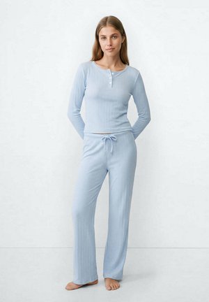 Young woman standing barefoot wearing light blue ribbed long-sleeve top with buttons and matching drawstring pants against white background.