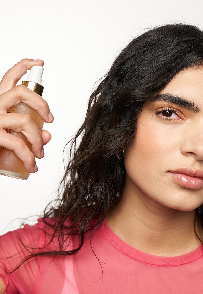 Klarer Sprühflasche mit goldener Kappe, die mit einer Hand gehalten wird, und einem hellbraunen Flüssigkeit darin. Die Person hat welliges dunkles Haar und trägt ein translucent rosa Oberteil.
